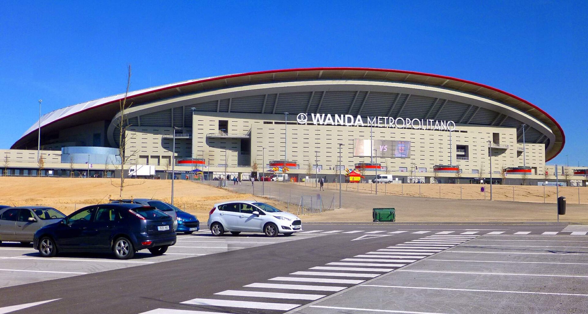 Football in Madrid: Bernabéu, Metropolitano and How to Get to a Match 3 Exterior of Atlético de Madrid's Metropolitano stadium with its curved roof edged in red, photographed across a car park under a clear blue sky