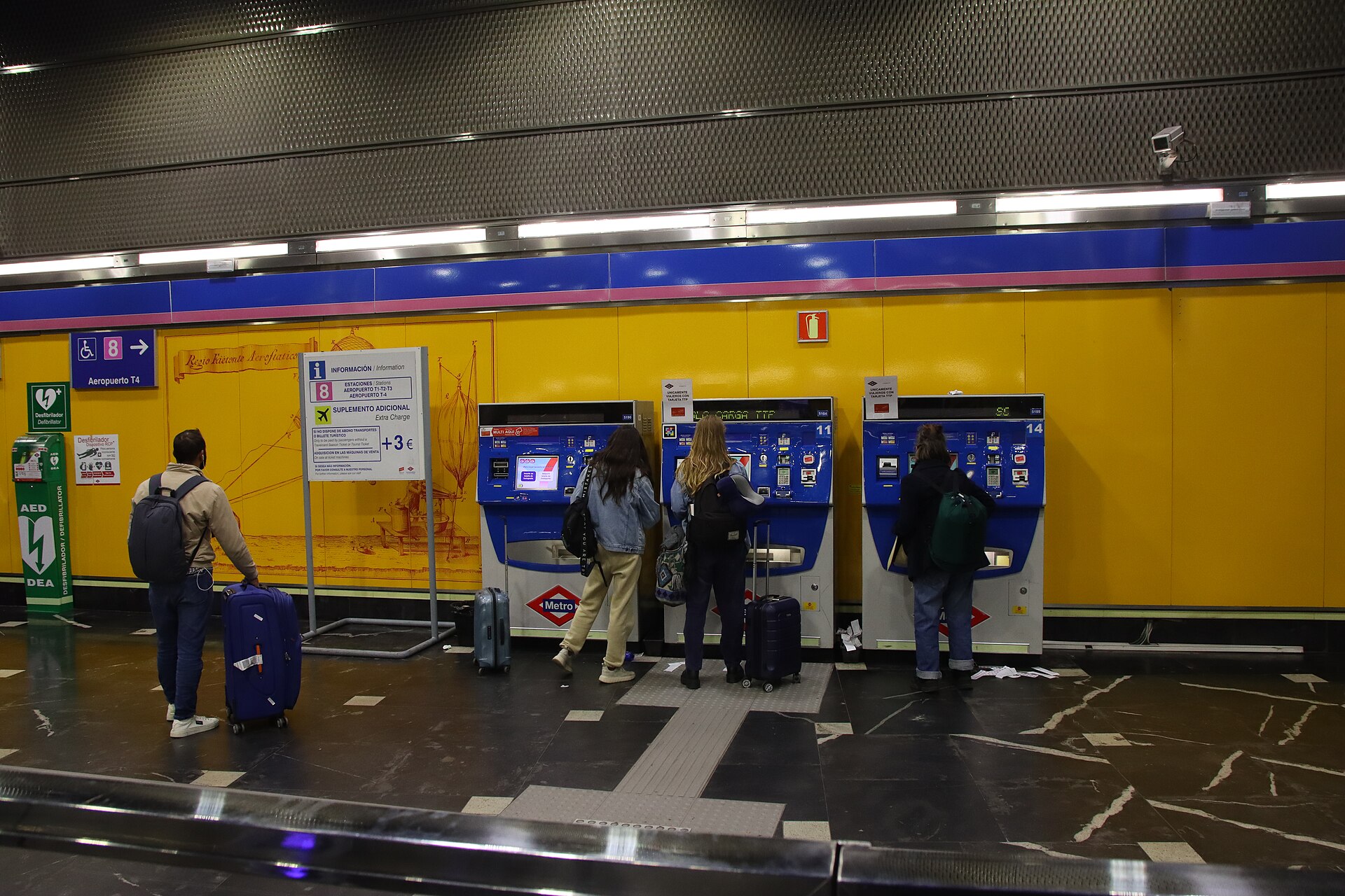 A Layover in Madrid: What to Do With 4, 6, 8 or 12 Hours 3 Travellers with suitcases buying tickets at blue Metro ticket machines in the Line 8 airport station, beside a sign showing the three-euro airport supplement