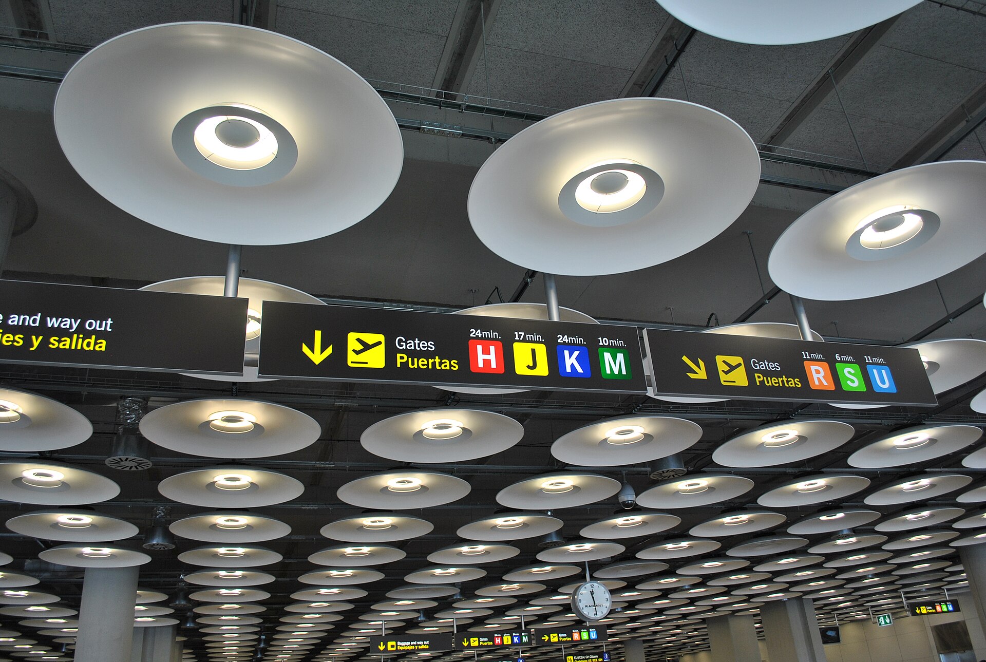 Overhead signs in Madrid Terminal 4 showing walking times to gates H, J, K and M and, on a second sign, to gates R, S and U, beneath round pendant lights