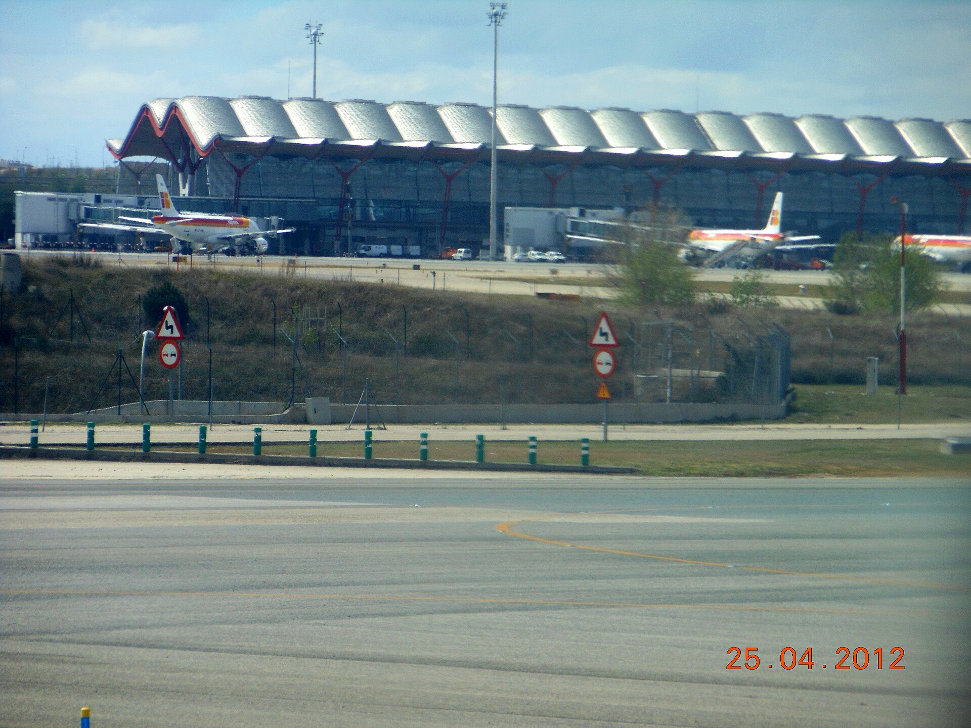 The long, wave-shaped roof of Madrid-Barajas Terminal 4 supported on red steel columns, with Iberia aircraft parked at the jet bridges in front