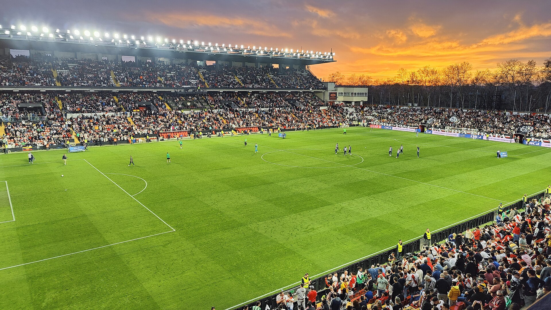 Football in Madrid: Bernabéu, Metropolitano and How to Get to a Match 4 Floodlit pitch at Rayo Vallecano's Estadio de Vallecas at half-time, with full stands and an orange sunset sky over the trees
