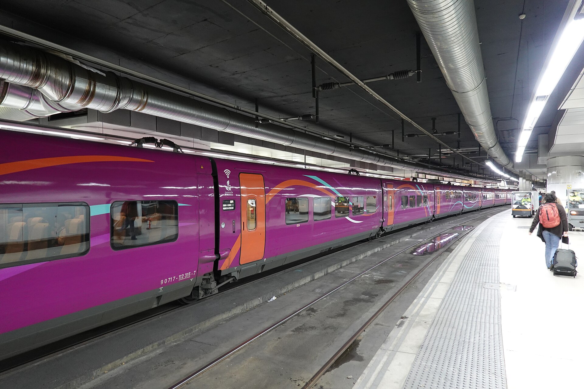 A purple and orange Avlo high-speed train standing at an underground platform with a passenger pulling a suitcase