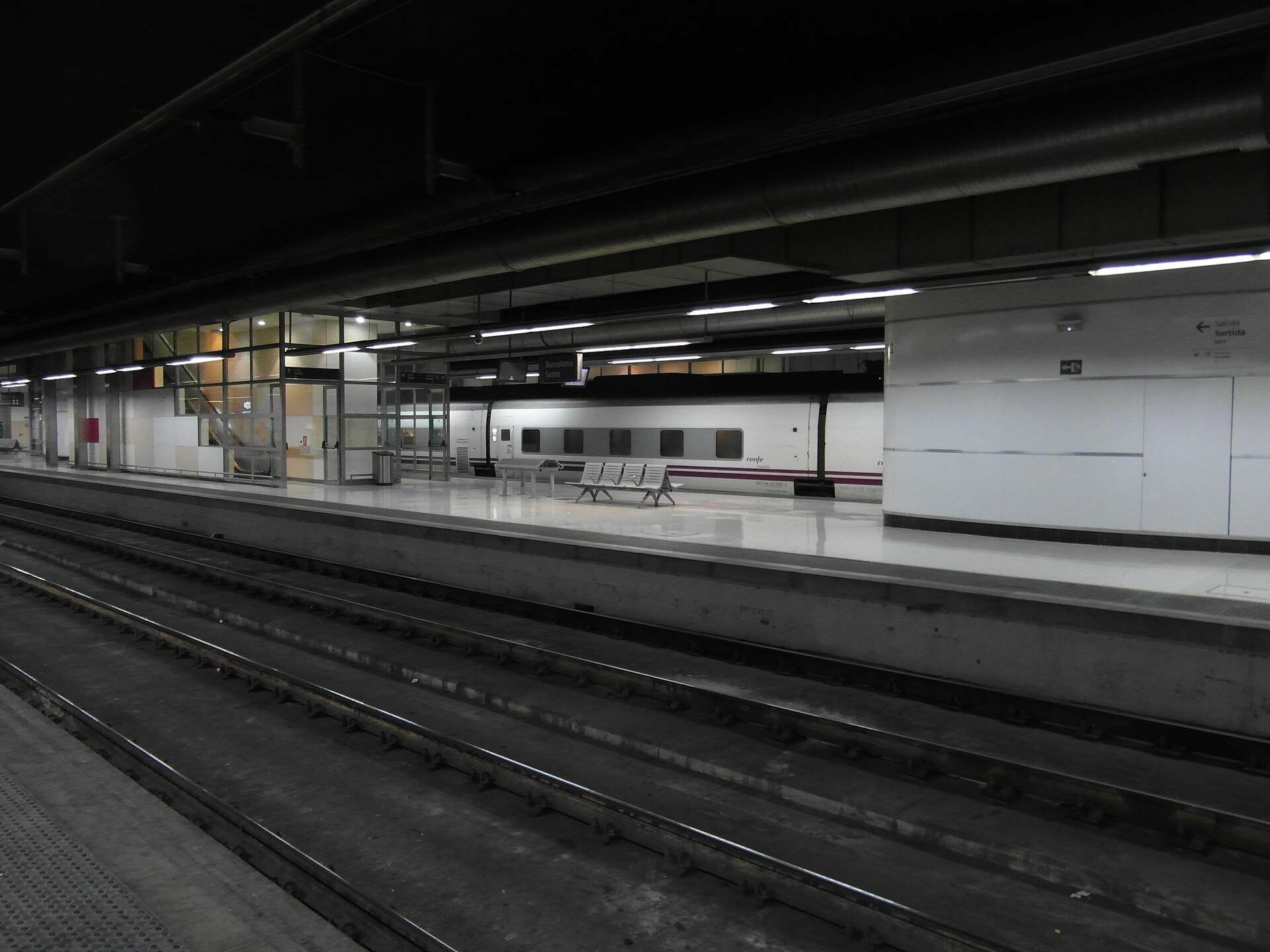 An underground platform at Barcelona Sants with a white Renfe train, empty benches and a Barcelona Sants sign