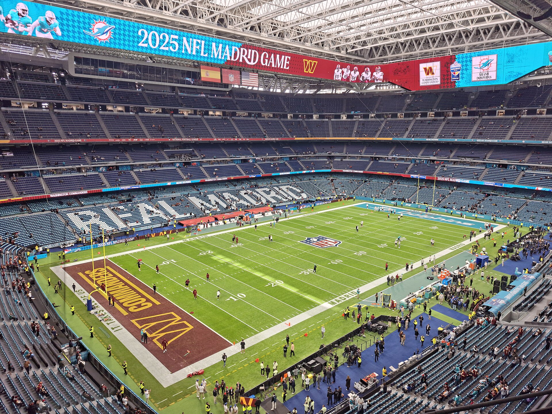 A stadium under a closed roof, its stands still mostly empty, with REAL MADRID spelled out in white seats in the lower tier behind a sports pitch
