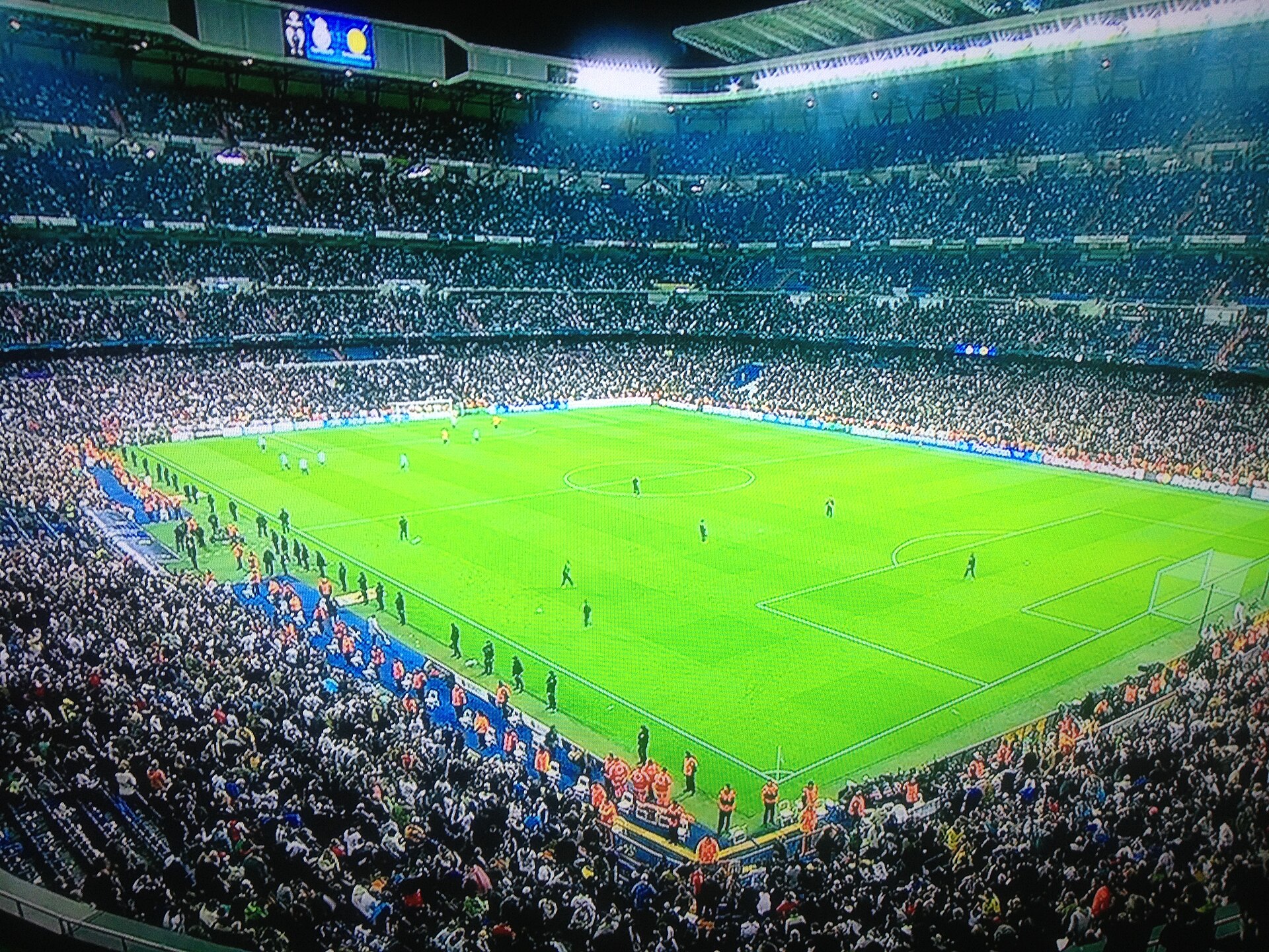 A football match under floodlights seen from high in a packed stadium, with the green pitch below and crowds on every side