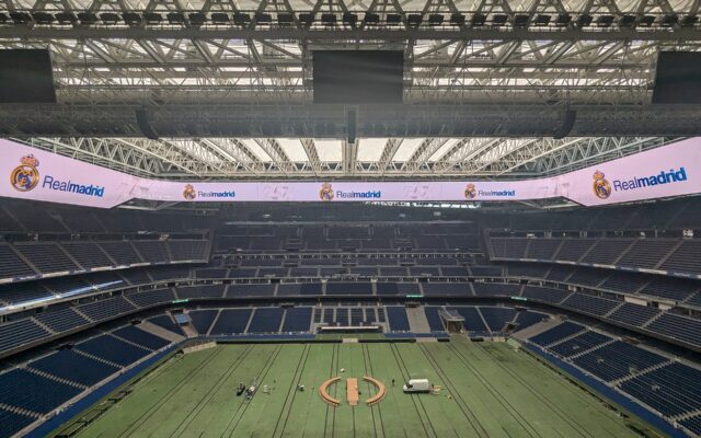 The inside of a large stadium with blue seats, a closed roof and a covered pitch