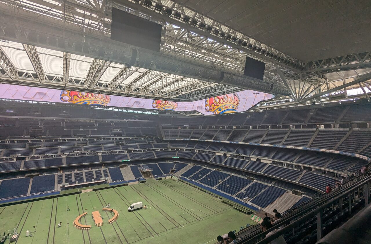The inside of a large stadium with blue seats, a covered pitch and a video screen running around the roof