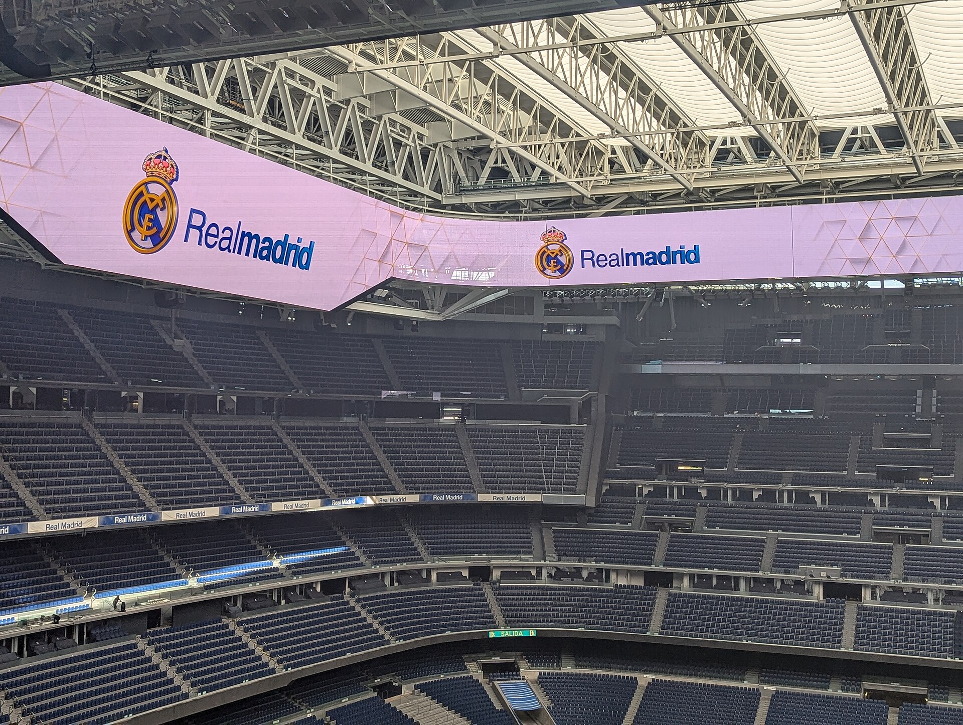 Empty blue seats rising in steep tiers, with Real Madrid crests on a continuous video screen under the roof