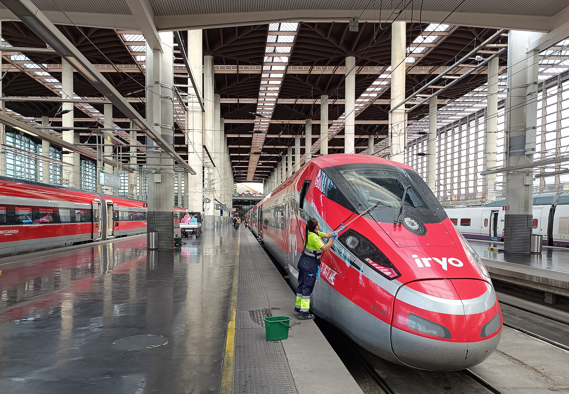 A cleaner washing the nose of a red iryo high-speed train beside a platform under a ribbed concrete and glass roof