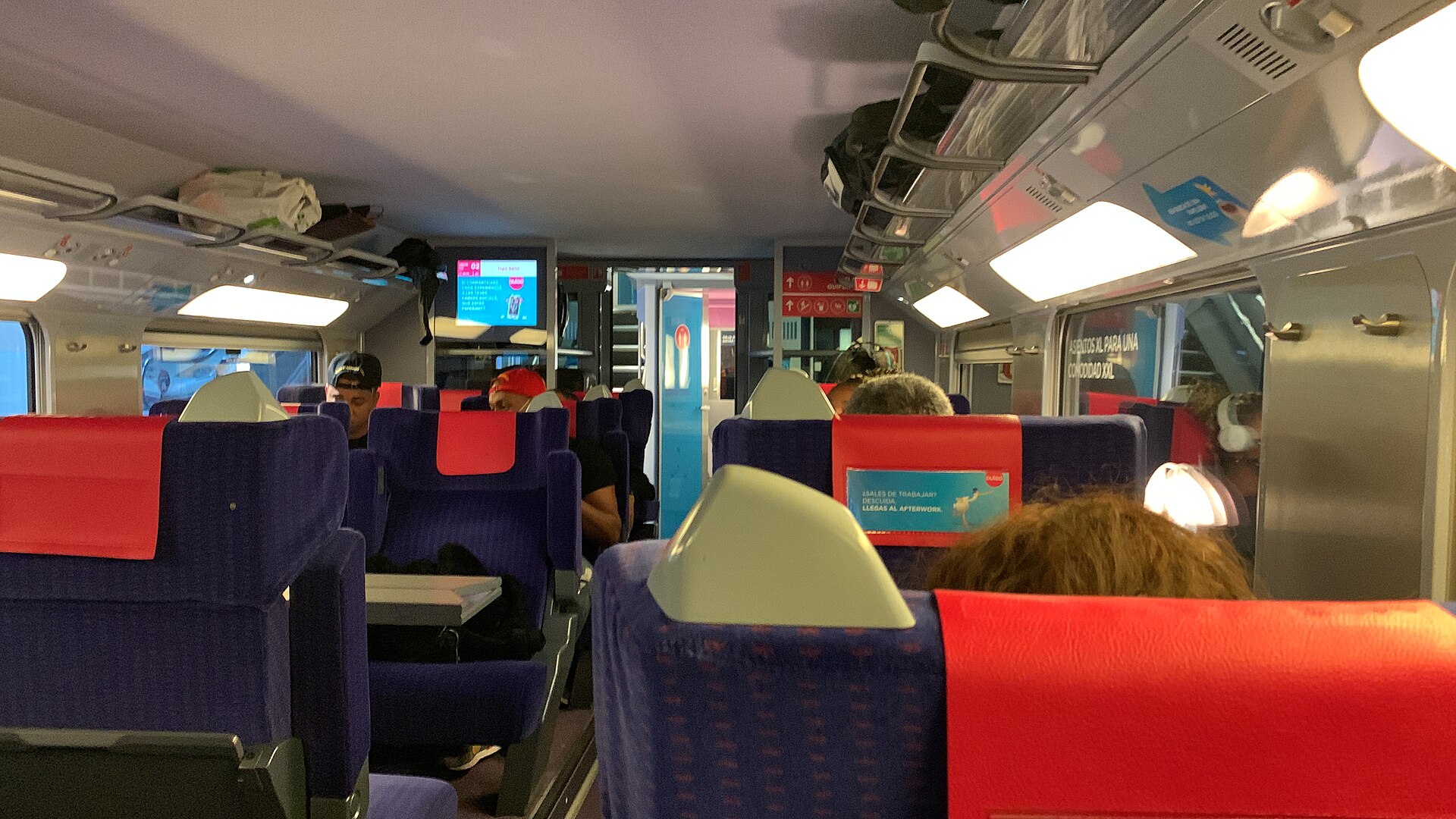 Blue seats with red headrest covers in a train carriage with overhead luggage racks and a screen at the far end