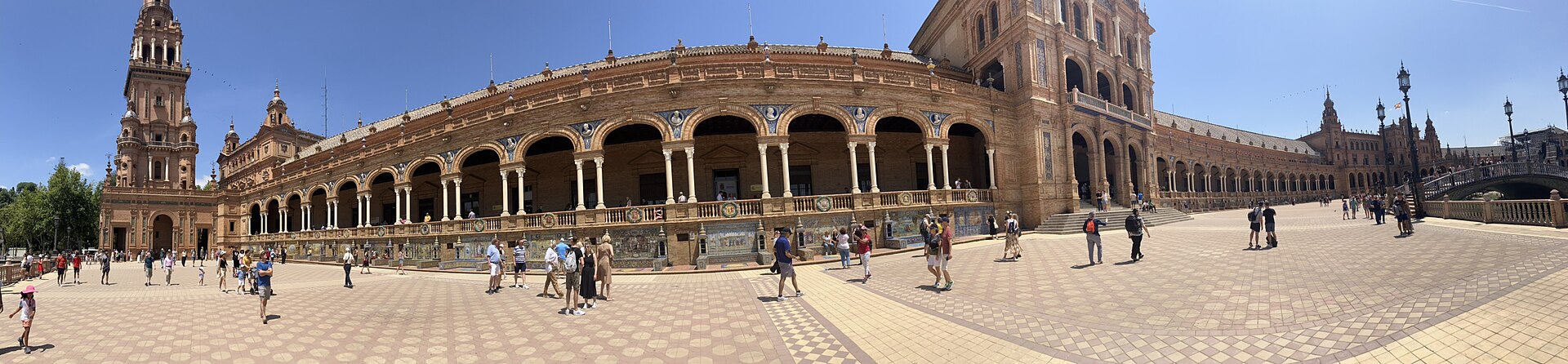 Panorama of a curved brick and tiled arcade with a tall tower at one end and visitors walking across a wide paved square