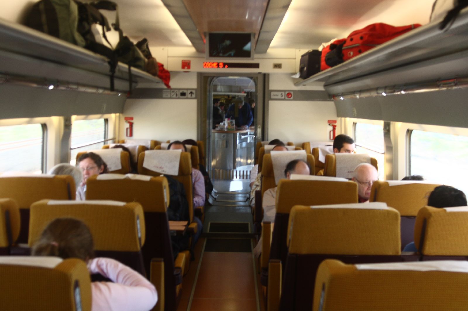 Rows of mustard-yellow seats with white headrest covers in a train carriage, with bags on the overhead racks