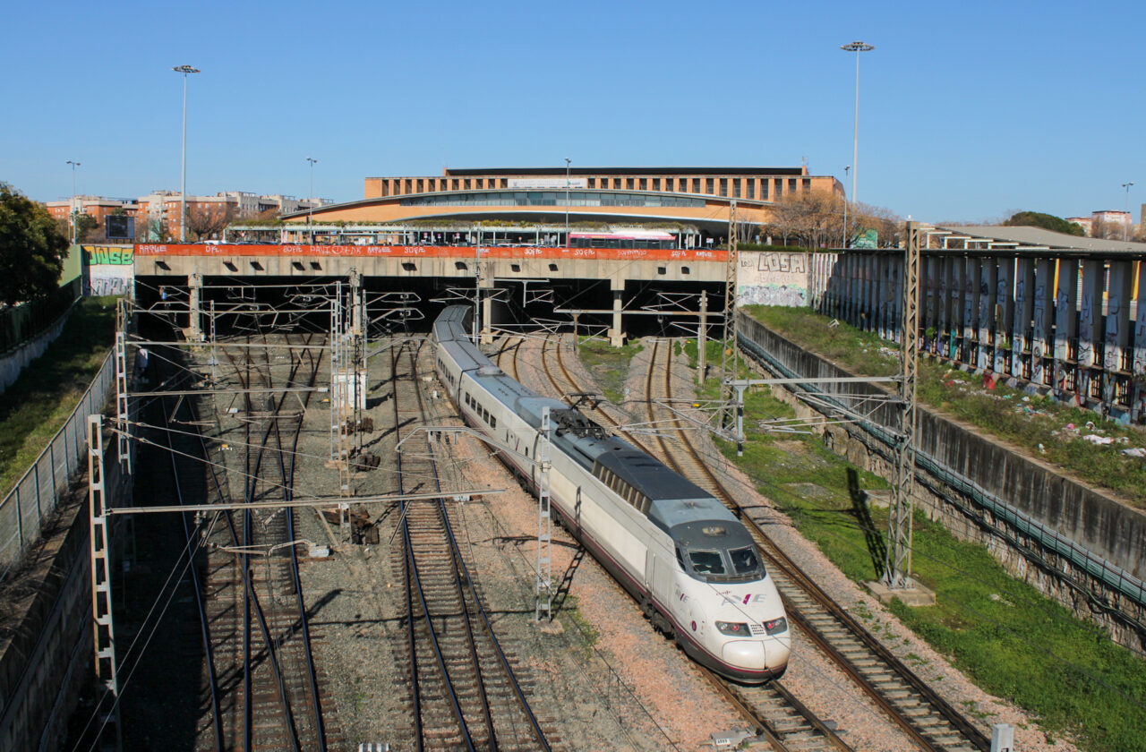 A white Renfe AVE train curving out from under a concrete deck in front of a long brick station building