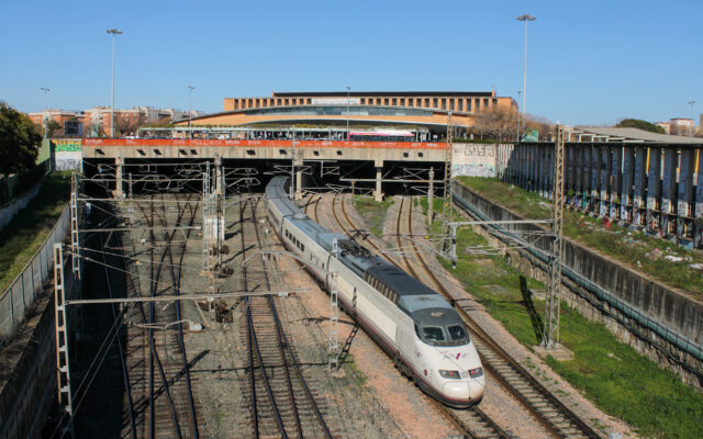 A white Renfe AVE train curving out from under a concrete deck in front of a long brick station building