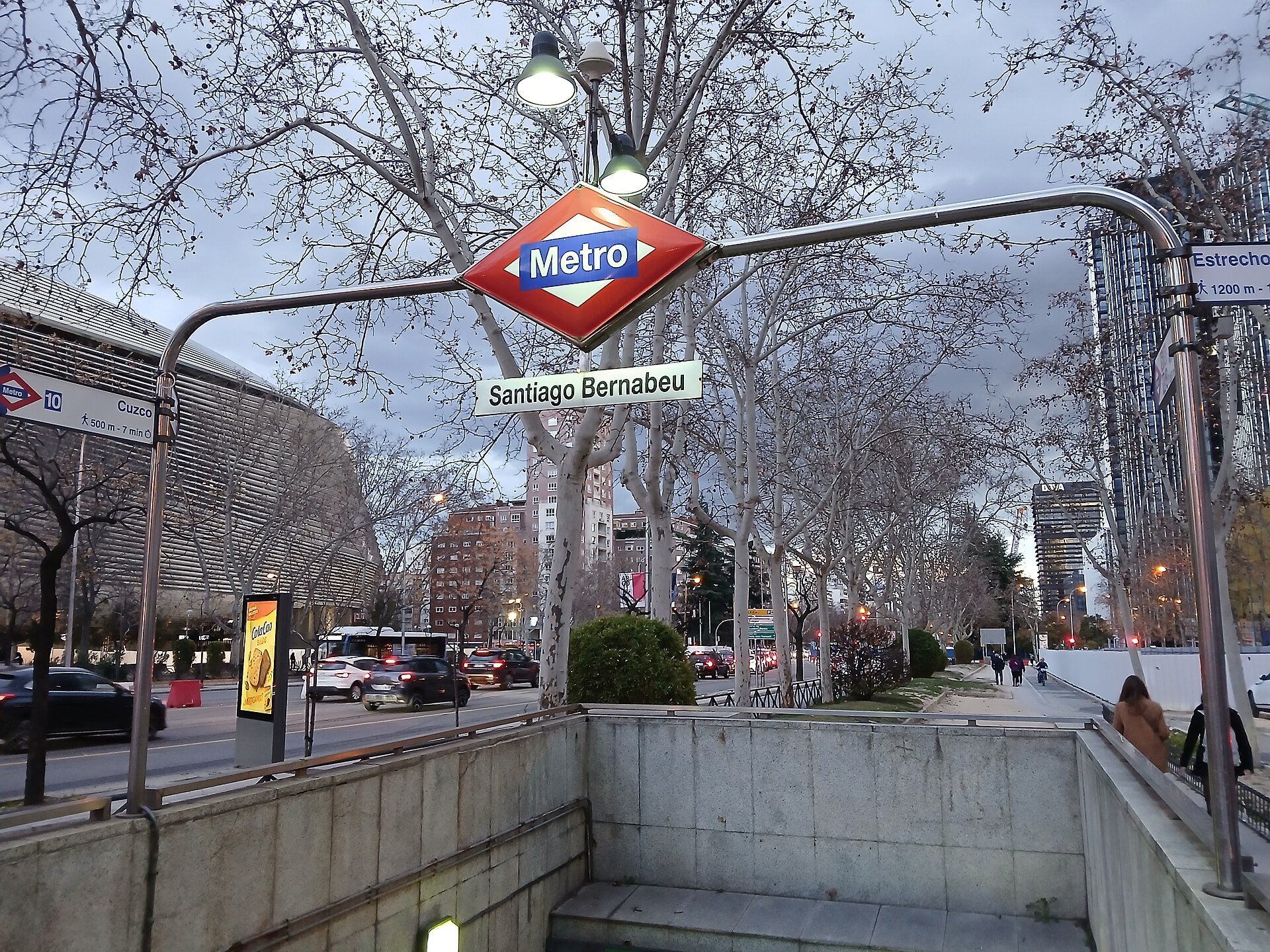 A red and blue Metro sign marked Santiago Bernabeu over a stairway entrance on a tree-lined avenue, with a large curved stadium facade behind