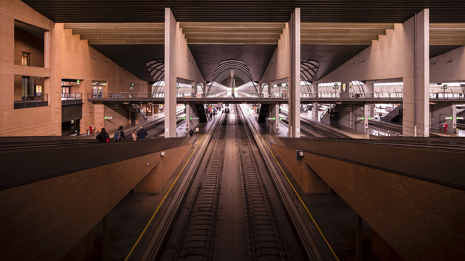 Looking down the tracks between two platforms inside a large brick and concrete station hall with a footbridge across it